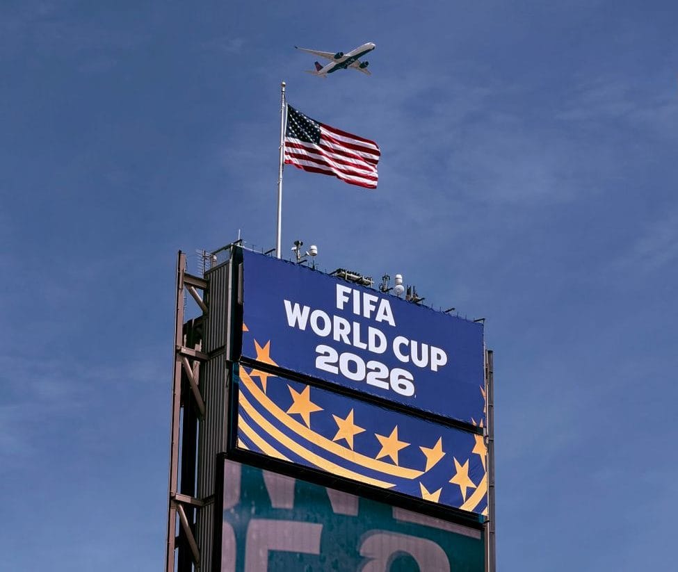 Airplane flying above FIFA World Cup 2026 sign with American flag, clear blue sky.